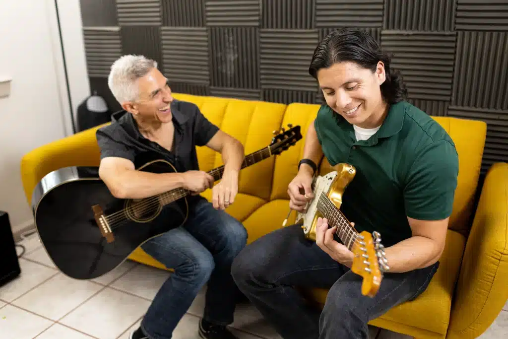 Instructor Bryan sits on a yellow couch with a student playing guitar at the Blue Frog School of Music, offering music lessons south Austin