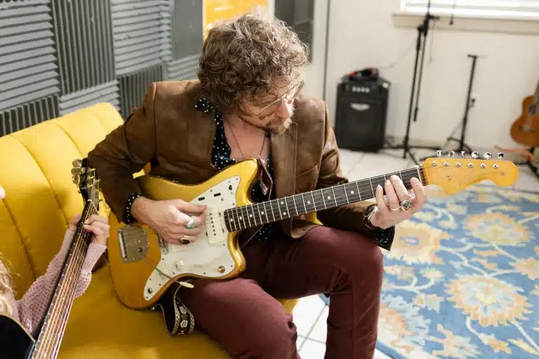 Instructor Nick sits on a yellow couch playing a gold electric guitar at the Blue Frog School of Music, offering music lessons south Austin