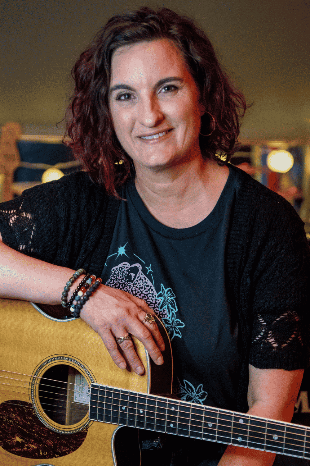 A photo of Kim Rae Andrulis holding a guitar and smiling at the camera at Blue Frog School of Music, where she is the Band Program Director in Oak Hill